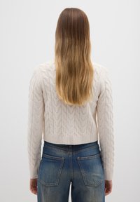 Gebreide trui in lichtbeige met een kabelpatroon, cropped ontwerp en geribbelde manchetten. Gecombineerd met hoog uitgesneden blauwe denim jeans.