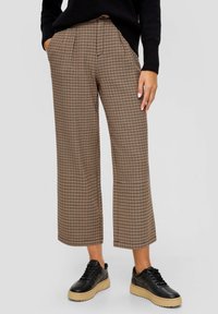 Pantalon large à plis avec motif pied-de-poule marron, porté avec des baskets plateformes noires. Le tissu présente une texture lisse.