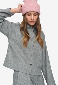 Eine Frau trägt eine pinke Strickmütze sowie eine passende graue strukturierte Jacke mit Knöpfen und Hose, während sie mit einer Hand den Hut richtet.