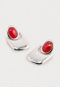 Srebrnasti naušnice s poliranim, apstraktnim oblikom i istaknutim crvenim ovalnim kamenom. Gladak premaz, moderan dizajn i jedinstvena silueta.