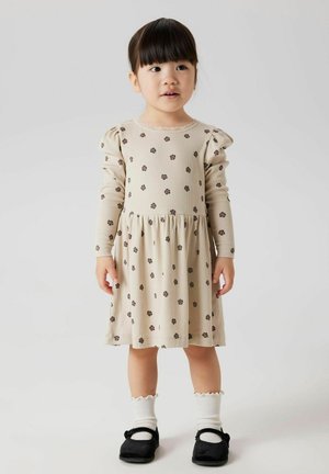 Robe beige à manches longues avec de petits motifs floraux sombres, texture côtelée, jupe froncée et col rond avec bordure en dentelle. Chaussures noires et chaussettes blanches.