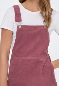 Femme portant une chemise blanche à manches courtes et une robe salopette en velours côtelé mauve avec des fermoirs métalliques et une main dans la poche.