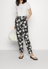 Pantalones estampados en blanco y negro con hojas de palma, camiseta blanca, bolso de hombro crema y sandalias negras con suela gruesa.