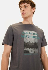 Grå bomulds t-shirt med et rektangulært grafisk motiv, der viser en naturscene, og teksten "Designet til hverdagens eventyr" i teal. Korte ærmer.
