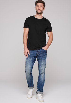 Black short-sleeve t-shirt, blue faded jeans, and white sneakers. Model stands with one hand in pocket against a neutral background.