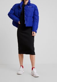 Veste d'extérieur bleue avec un col haut et des boutons-pression, associée à une robe midi noire à côtes et des baskets blanches avec des accents rouges.