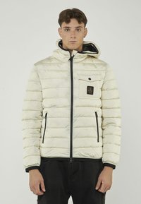 Giacca imbottita di colore crema con chiusura a zip, dettagli neri e una tasca frontale con un patch del logo, design trapuntato testurizzato.