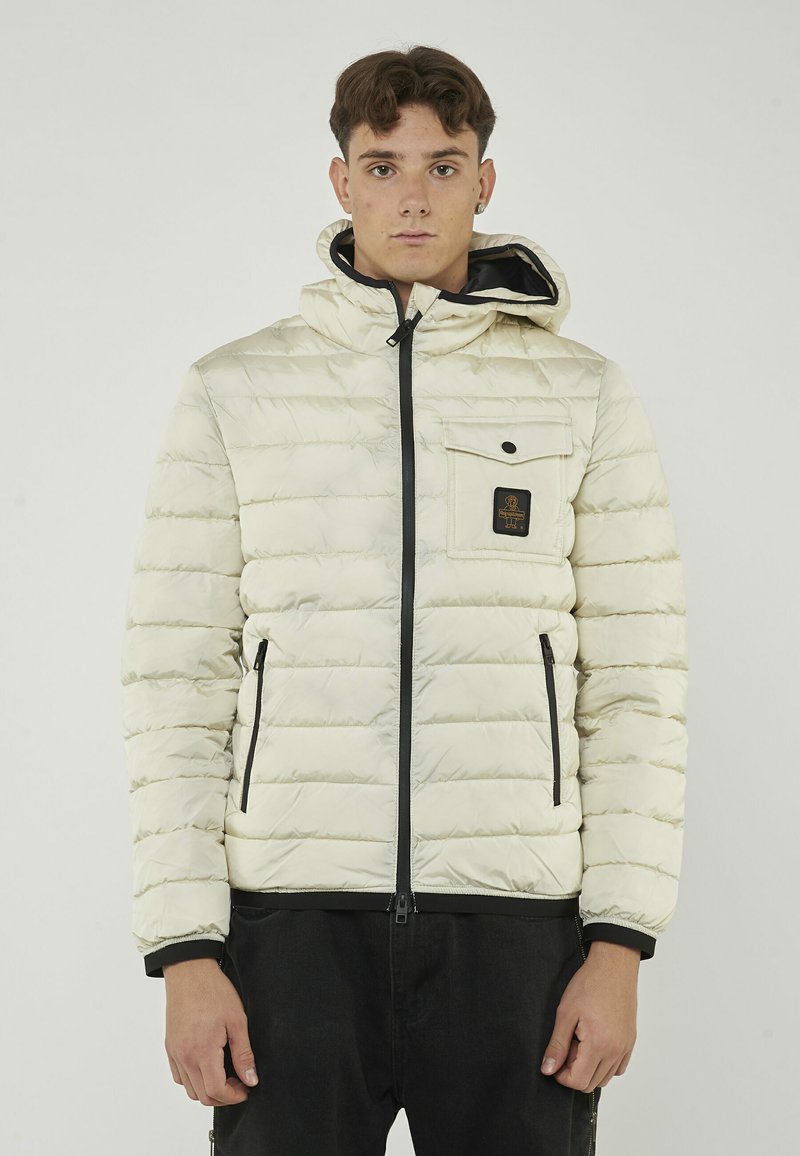 Giacca imbottita di colore crema con chiusura a zip, dettagli neri e una tasca frontale con un patch del logo, design trapuntato testurizzato.