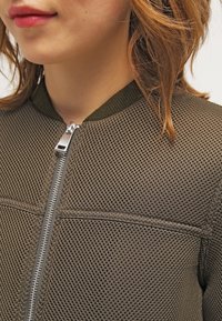 Oliwkowa siateczkowa kurtka bomber z srebrnym zamkiem błyskawicznym, z prążkowanym kołnierzem i teksturowanym materiałem, z detalami przeszyć i panelowymi sekcjami.