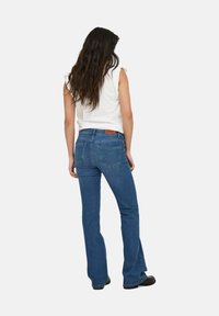 Jeans acampanados azules de mezclilla, con dos bolsillos traseros y una etiqueta de cuero marrón en la cintura. Combinados con una blusa sin mangas blanca.