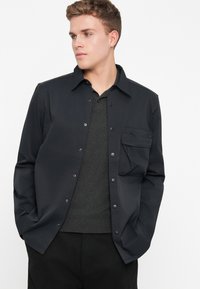 Zwarte button-up overhemd met een borstzak, gemaakt van gladde stof. Kenmerkt zich door een klassieke kraag en een eenvoudig ontwerp, gelaagd over een donkergrijze trui.