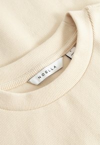 Gros plan sur un tissu côtelé de couleur crème montrant un col rond et une étiquette de vêtement blanche avec le texte "NOELLA".