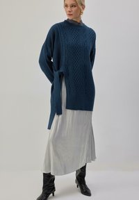 Pull tricot surdimensionné bleu sarcelle avec motifs texturés et liens latéraux, associé à une jupe blanche plissée fluide et des bottes noires montant jusqu'aux genoux.