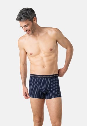 Homme torse nu souriant, portant un boxer bleu marine à motif de points blancs et une ceinture de marque « Eminence », debout devant un fond uni.