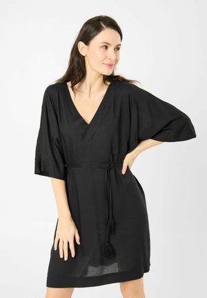 Frau trägt ein knielanges schwarzes Kleid mit weiten Ärmeln und einem Quasten-Gürtel, steht mit einer Hand in der Hüfte und schaut seitlich.