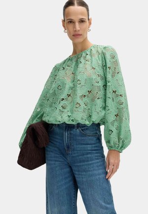 Kvinde iført en grøn blomstret blondebluse med lange, puffede ærmer, blå jeans og holder et sammenfoldet mørkebrunt tekstureret stofaccessory.