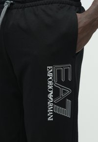 Μαύρο φόρμας από απαλό ύφασμα· διαθέτει λευκό τυπωμένο λογότυπο "EA7 EMPORIO ARMANI" κατά μήκος της πλευράς, με ελαστική μέση και κορδόνι.