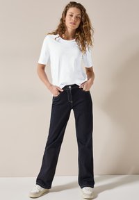 Jeans blu scuro a gamba larga con cuciture gialle a contrasto, abbinati a una maglietta bianca a maniche corte semplice. Sneakers bianche visibili in basso.