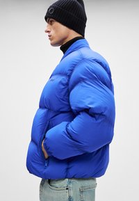 Mėlynas pūkinis džemperis su blizgiu paviršiumi, su užtrauktukais kišenėse, raukšlėtomis rankovėmis ir dėžutės siluetu, dėvimas ant juodo golfuko ir šviesių džinsų.