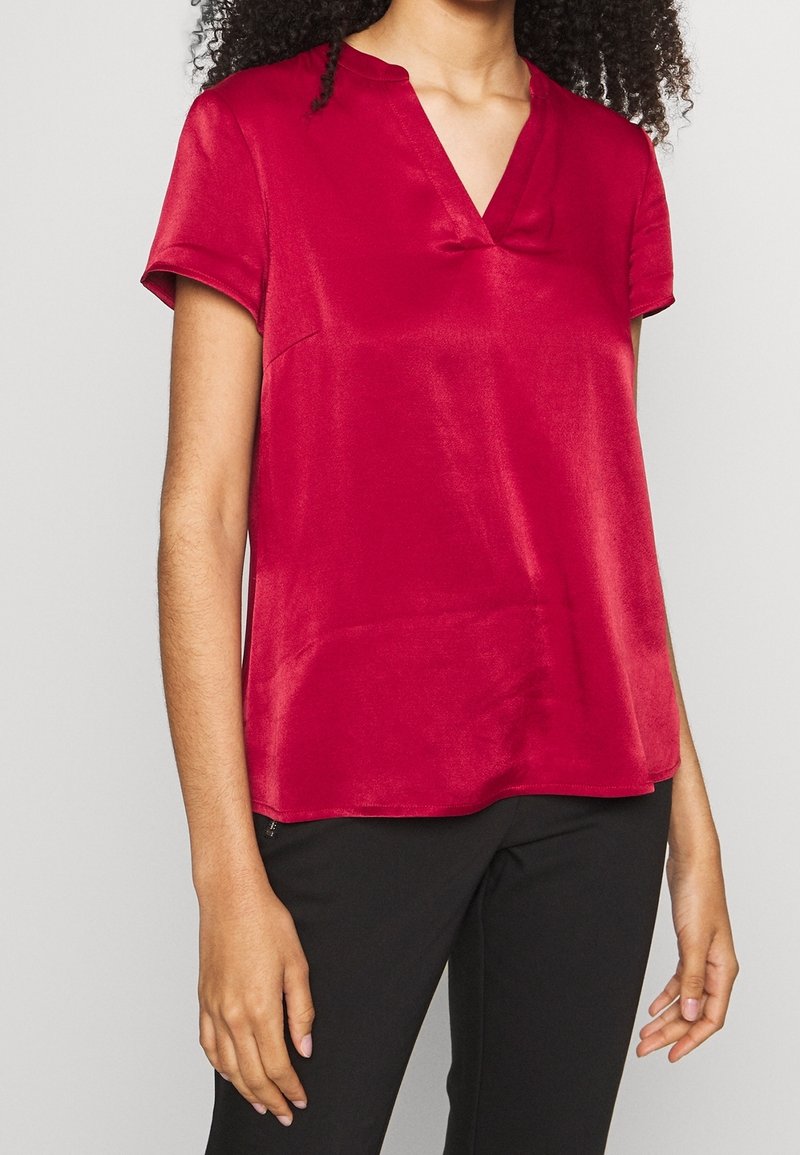Femme portant un chemisier rouge brillant à manches courtes avec un col en V et un pantalon noir, debout devant un fond clair et uni.