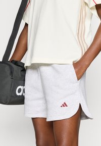 Svetlo sive bombažne kratke hlače z bočnimi razporki in bordo Adidas logotipom. Kombinirane s kremnim topom z kontrastnimi vertikalnimi črtami.