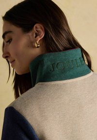 Pletený sveter s béžovým telom, zeleným golierom a námorníckymi rukávmi. Na golieri je vyšitý nápis "JOULES" a materiál má textúrovanú štruktúru.