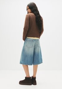 Pulover maro, cu tiv galben deschis, culottes din denim albastru cu un finisaj uzat și bocanci scurți maro închis. Material texturat și croială lejeră.