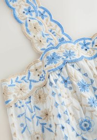 Tissu blanc avec broderie florale bleue et beige, présentant des bords festonnés et un détail de conception froncée.