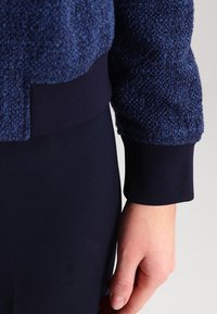 Dunkelblauer strukturierter Pullover aus weichem, geschlungenem Material am Körper und glatten, taillierten Bündchen. Nahaufnahme von Ärmel und Hand.
