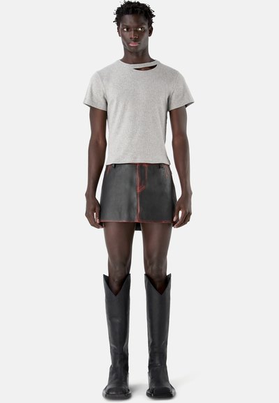 Modello maschile che indossa una t-shirt grigia corta con un taglio, minigonna in pelle nera con cuciture rosse e stivali neri alti.