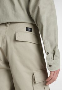Beige cargo pants made of cotton with a patch pocket and black label. Light green long-sleeve shirt with a ribbed texture and buttoned cuffs.