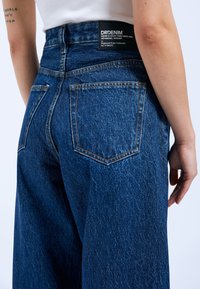 Dunkelblaue, weit geschnittene Jeans aus Denim, mit gelben Nähten, zwei Gesäßtaschen und einem schwarzen Etikett mit Markeninformationen am Bund.