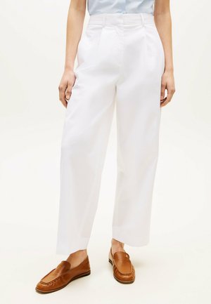 Mujer con pantalones blancos de tiro alto y pata ancha y mocasines de cuero marrón, de pie contra un fondo liso y claro.