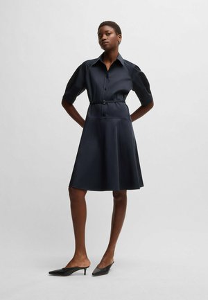 Navy shirt dress with short sleeves, belted waist, and flared skirt. Smooth, glossy fabric. Paired with black, pointed toe mules.