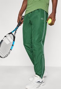 Pantalones deportivos verdes con rayas blancas a los lados, hechos de un material suave. Se sostiene una raqueta de tenis en una mano y una pelota de tenis en la otra.