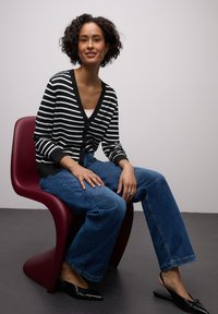 Sort-hvid stribet cardigan med knapper og v-udskæring, parret med brede blå jeans. Modellen sidder på en bordeauxfarvet stol.