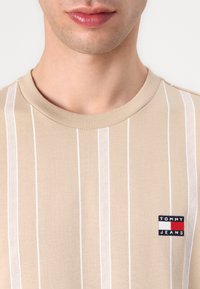 Beige T-shirt with white vertical stripes, round neck, and small "Tommy Jeans" logo patch in the lower left corner. Smooth texture.
