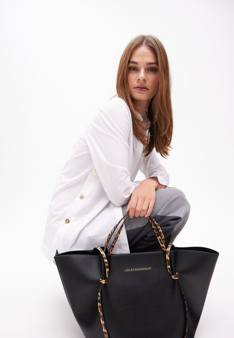 A young woman in a white button-up shirt and gray pants squats, holding a black handbag with leopard-print straps, gazing confidently.