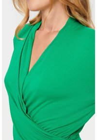 Grön omlottklänning med djup V-krage, mjuk textur och draperat tyg som skapar mjuka veck längs bysten.