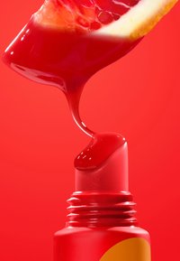 Czerwony błyszczyk w cylindrycznej tubce z fakturowaną nakrętką; błyszcząca ciecz spływa z aplikatora, podkreślona plasterkiem pomarańczy.