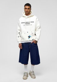 Witte hoodie met zwarte tekst en verfspetters accentueert de denim shorts. Het model draagt witte sokken en grijze sneakers.