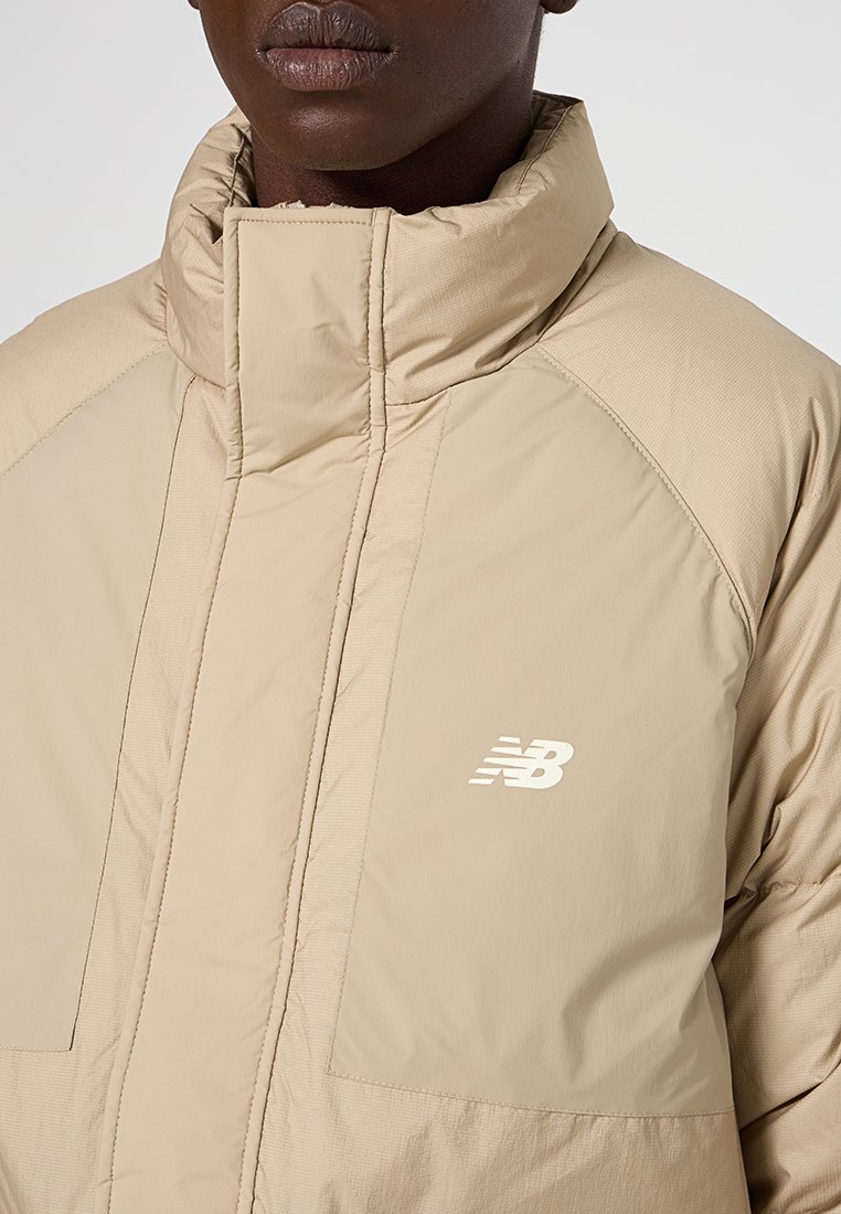 Chaqueta acolchada beige con cuello alto, cierre de cremallera en la parte delantera y bolsillos de parche. Presenta un logo blanco incrustado en el pecho y una textura suave y acolchada.