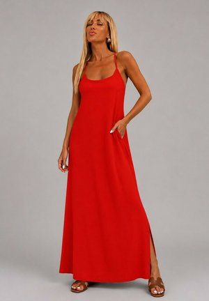 Femme portant une longue robe rouge sans manches avec de fines bretelles, une fente latérale et des sandales marron, debout devant un fond gris uni.