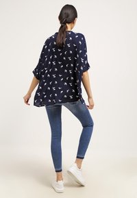 Blouse bleu marine avec motif d'oiseaux blancs, coupe ample, manches courtes et ourlet arrondi, associée à un jean skinny bleu clair et des baskets blanches.