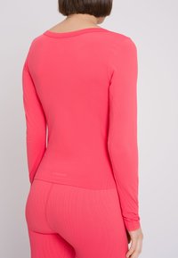 Maglietta sportiva a maniche lunghe in tessuto rosa brillante, con scollatura a girocollo e motivi testurizzati sui lati, abbinata a leggings rosa coordinati.
