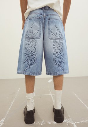 Osoba v svetlomodrých rifľových šortkách s čiernou kresbou kmeňového plameňa na zadných vreckách a nohaviciach, biele ponožky a čierne topánky.