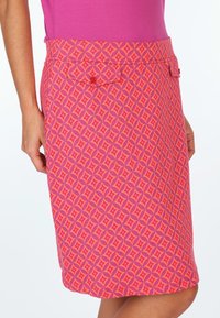 Pink skirt featuring a geometric pattern in lighter pink and orange hues, with a back pocket and button detail. Smooth, textured fabric.