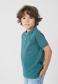 Jong kind met krullend bruin haar, gekleed in een teal polo en lichte spijkerbroek, staande met één hand in de zak tegen een effen achtergrond.