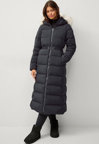 Manteau long noir matelassé avec capuche en fourrure, taille cintrée, fermeture éclair complète et poches passepoilées. Porté avec des leggings noirs et des bottines.