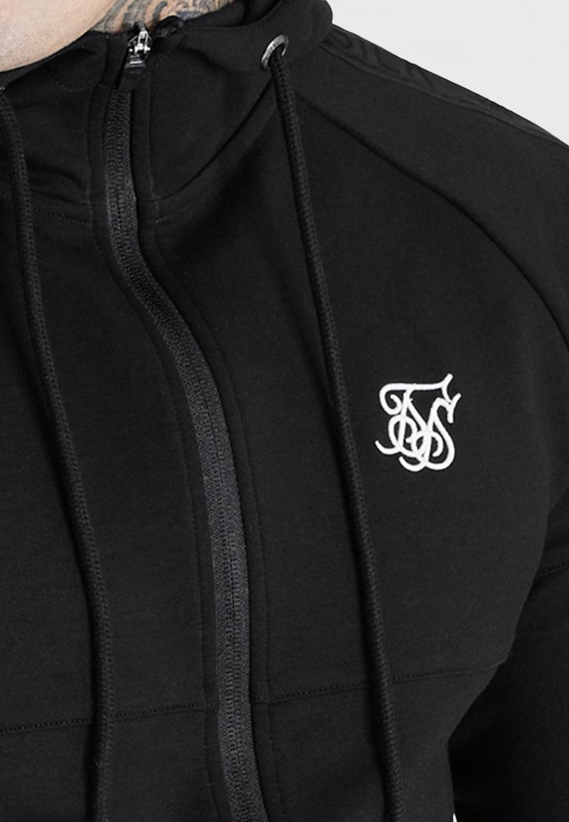 Sweat à capuche zippé noir avec cordons et logo brodé blanc sur la poitrine représentant des lettres stylisées.